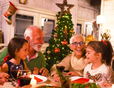 Family celebrating Christmas dinner with two children, an older man, and a woman by a decorated tree.