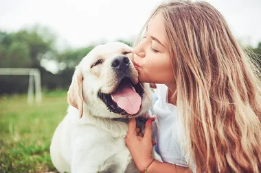 Young woman kissing a happy Labrador retriever outdoors on a grassy field.