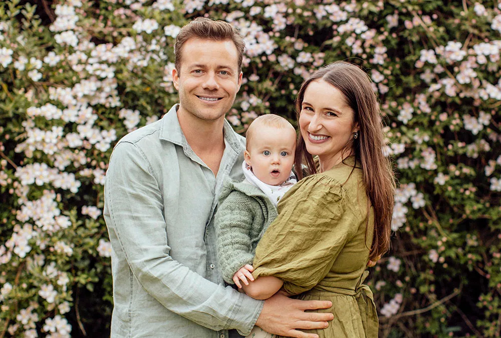 A smiling couple holds a baby in front of flowering bushes; all are wearing green.