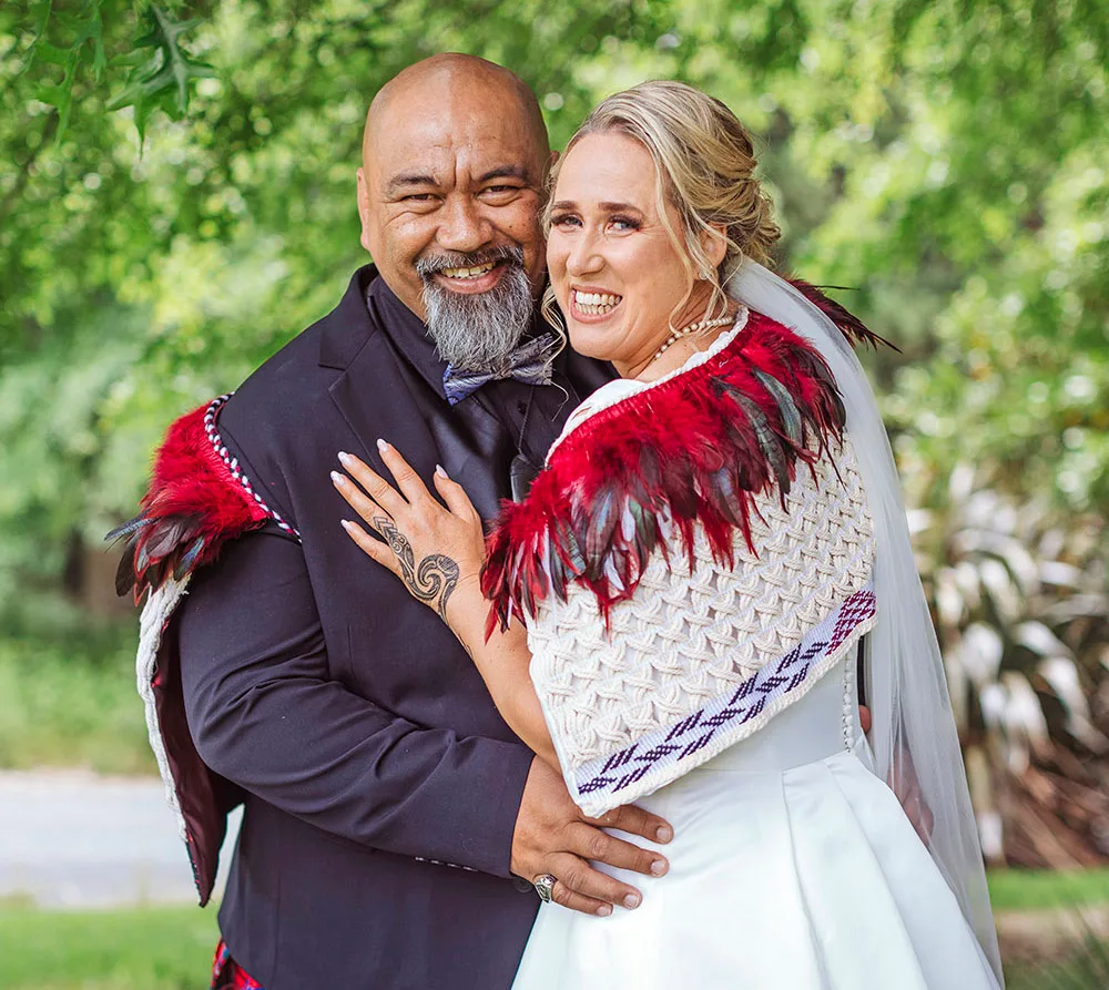 A couple in traditional Māori attire embraces outdoors, smiling broadly under a sunny, leafy background.