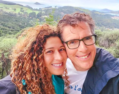 Two people smiling in a selfie with lush green hills and the ocean in the background.