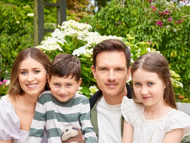 Family of four smiling in a garden, with two children holding a stuffed animal, surrounded by flowers and greenery.