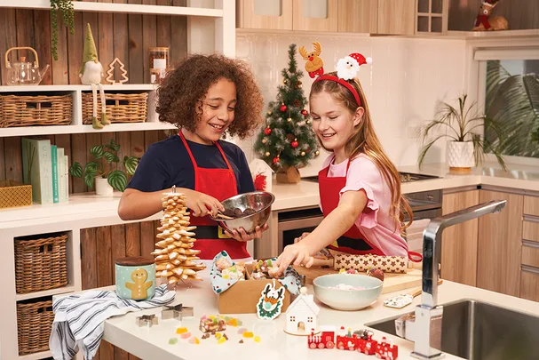 Two children in a kitchen making holiday cookies with decorations, wearing festive headbands and red aprons.