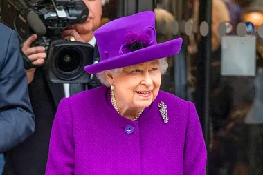 A person in a vibrant purple outfit and hat, smiling, with a camera crew in the background.
