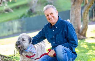 Man kneeling and smiling next to a fluffy dog wearing a bandana, in a sunny park setting.