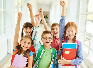 Five smiling children with raised arms holding books and backpacks in a brightly lit hallway.