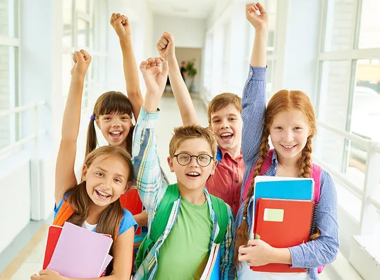 Five smiling children with raised arms holding books and backpacks in a brightly lit hallway.