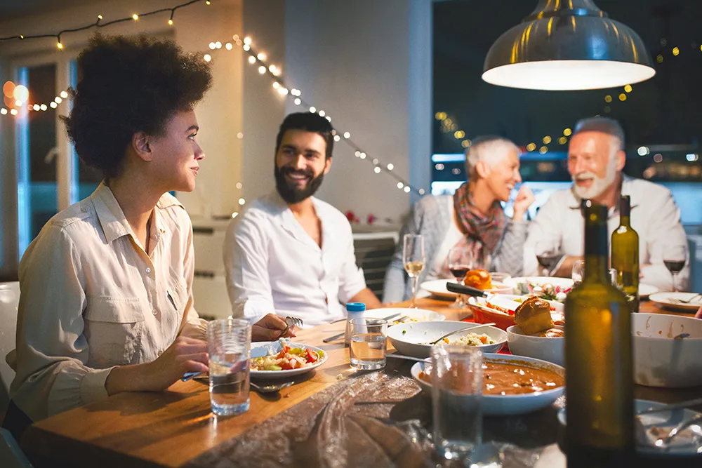 A group of people enjoying a festive dinner at a warmly lit table, with food and drinks, and a backdrop of string lights.