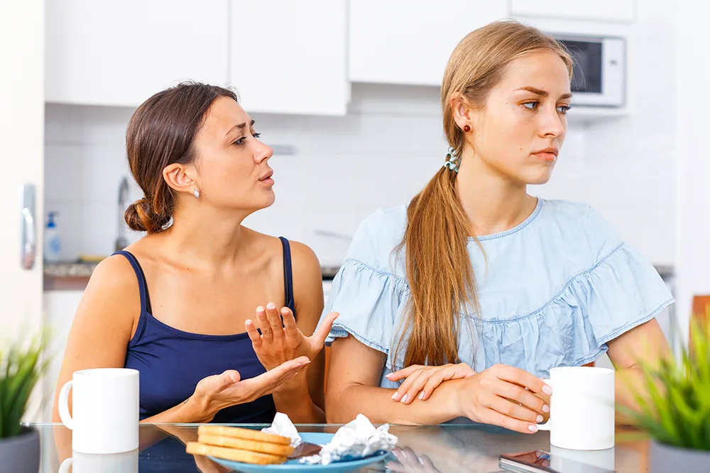Two women sitting at a table in a kitchen, one looking upset while the other gestures and talks.