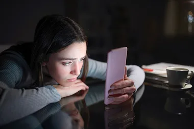 Young woman in dimly lit room, focused on her phone, with a cup and papers on the table.