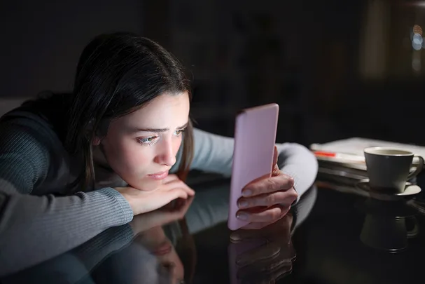 Young woman in dimly lit room, focused on her phone, with a cup and papers on the table.