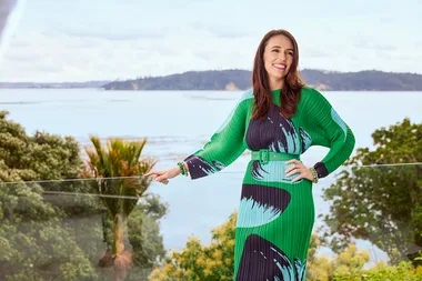 A woman in a green patterned dress stands on a balcony with a scenic view of water and trees in the background.