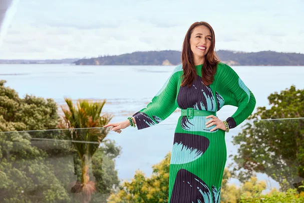 A woman in a green patterned dress stands on a balcony with a scenic view of water and trees in the background.