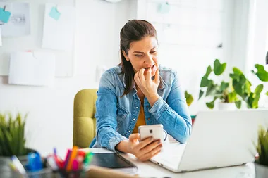 Woman biting nails, looking at phone, sitting at desk with laptop; appears worried or anxious in home office setting.