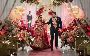 Indian bride in red lehenga and groom in tuxedo walking down a flower-adorned aisle with large floral arrangements.