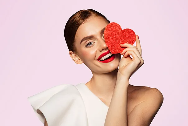 Woman with red lips smiling, holding a red glitter heart over one eye, against a soft pink background.