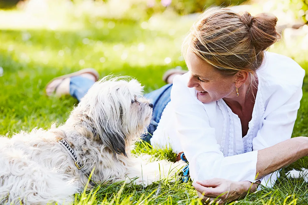 A woman lying on grass, smiling at her fluffy dog, who is looking back at her.