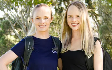 Two women smiling outdoors, wearing backpacks, surrounded by trees.