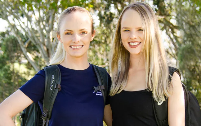 Two women smiling outdoors, wearing backpacks, surrounded by trees.