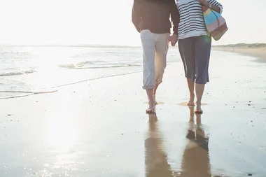 Couple walking hand in hand on a sunny beach, barefoot, with reflections in wet sand.
