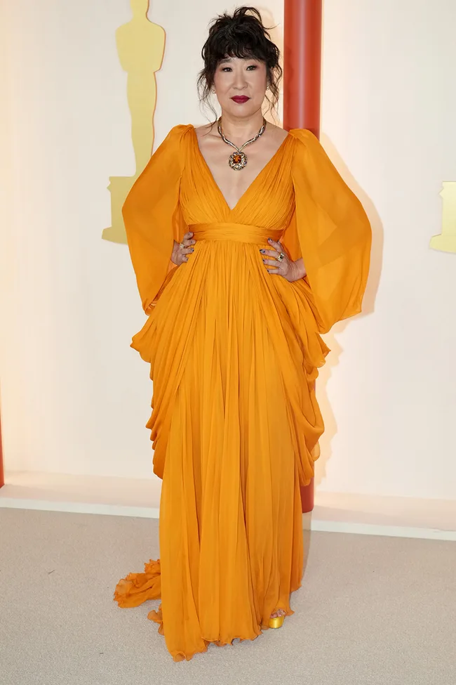 Person in an orange gown poses at an event with an Oscar statue in the background.