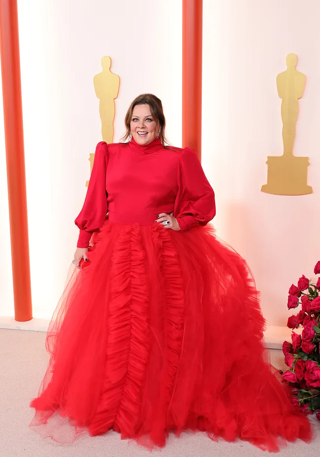 Person in a vibrant red gown stands smiling against an Oscars-themed backdrop.