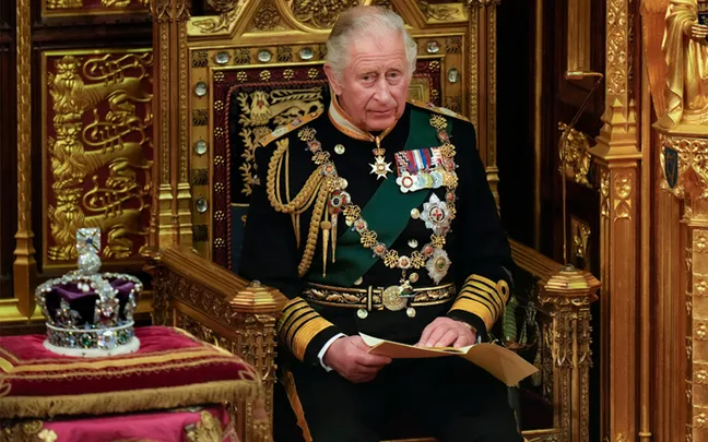 Man in ornate military uniform seated on a throne, holding papers, with a jeweled crown placed nearby.
