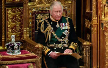 Man in ornate military uniform seated on a throne, holding papers, with a jeweled crown placed nearby.