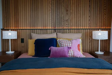 Bed with colorful pillows, wooden panel background, two white lamps on bedside tables, and soft ambient lighting.