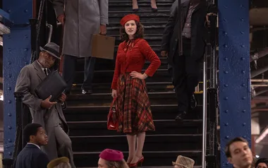Woman in red jacket and plaid skirt stands confidently on a staircase in a busy subway station.