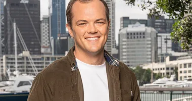 A smiling man in a brown jacket and white shirt with an urban marina background.