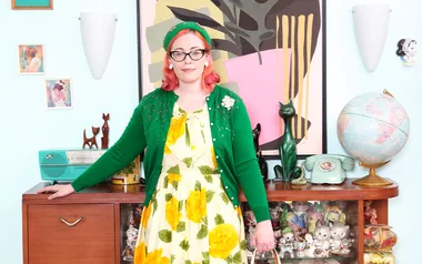 Woman in vintage floral dress and green cardigan stands in retro-styled room with globe, phone, and decor items.
