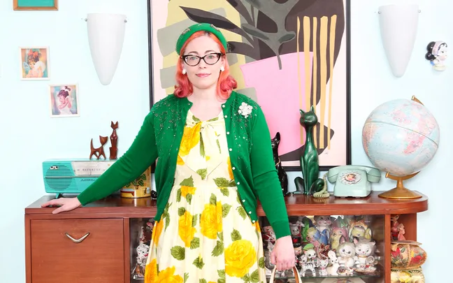 Woman in vintage floral dress and green cardigan stands in retro-styled room with globe, phone, and decor items.