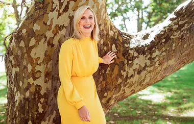 A woman in a bright yellow dress smiles while leaning against a large tree in a sunlit park.