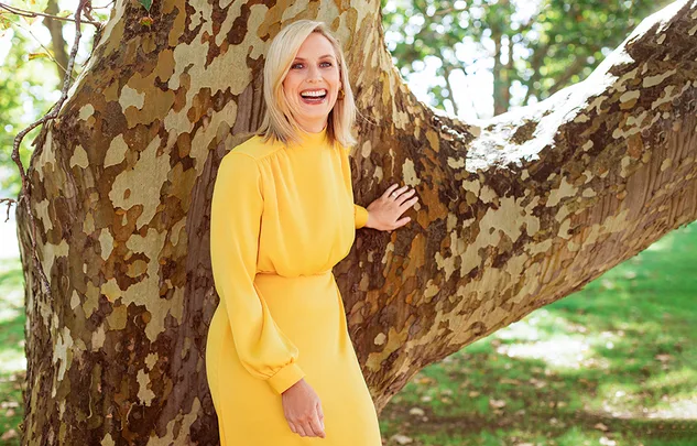 A woman in a bright yellow dress smiles while leaning against a large tree in a sunlit park.