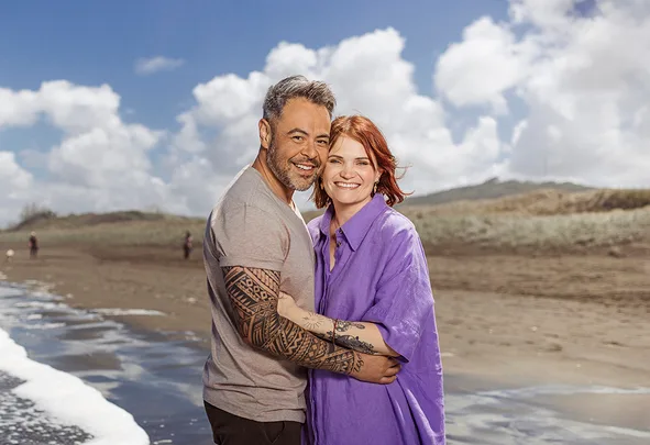 Couple hugging on a beach, smiling, with waves and cloudy sky in the background.