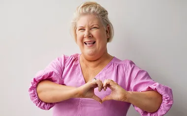 A woman in a pink blouse makes a heart shape with her hands, smiling brightly against a light background.