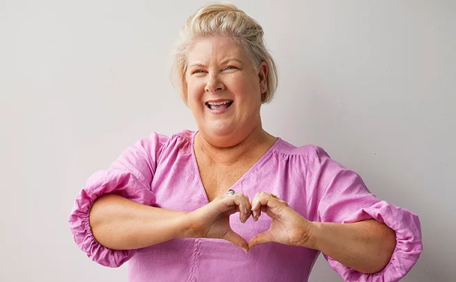 A woman in a pink blouse makes a heart shape with her hands, smiling brightly against a light background.