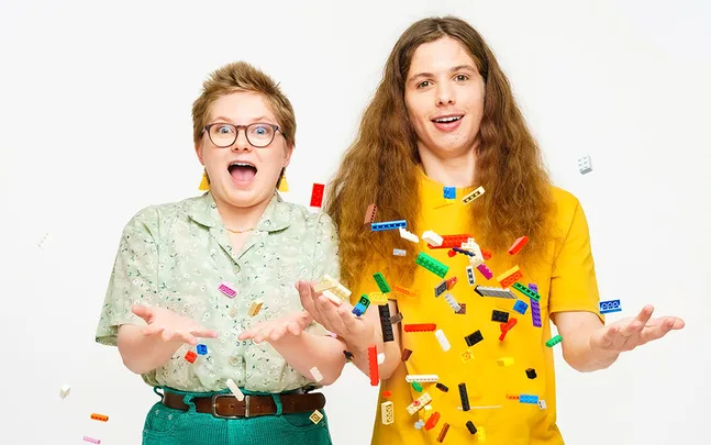 Two people smiling while colorful LEGO bricks float in the air around them.