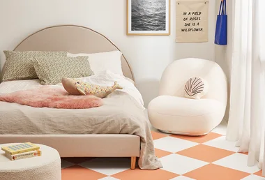 Cozy bedroom with a beige bed, floral pillows, plush chair, wall art, and a soft pink rug on a checkered floor.