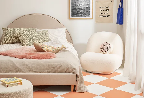 Cozy bedroom with a beige bed, floral pillows, plush chair, wall art, and a soft pink rug on a checkered floor.