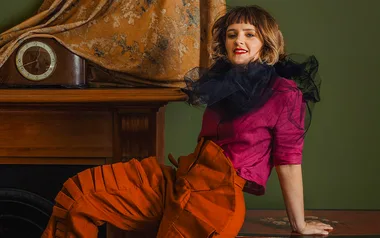 A person in a colorful outfit and black scarf poses confidently in a vintage room with a clock on the mantel.