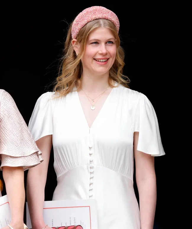 A woman in a white dress with a pink headband, holding documents, smiles against a dark background.