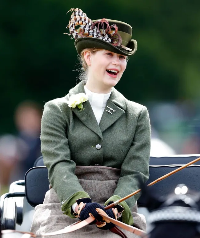 A person wearing a green hat with feathers and a matching jacket, smiling while holding reins, outdoors.