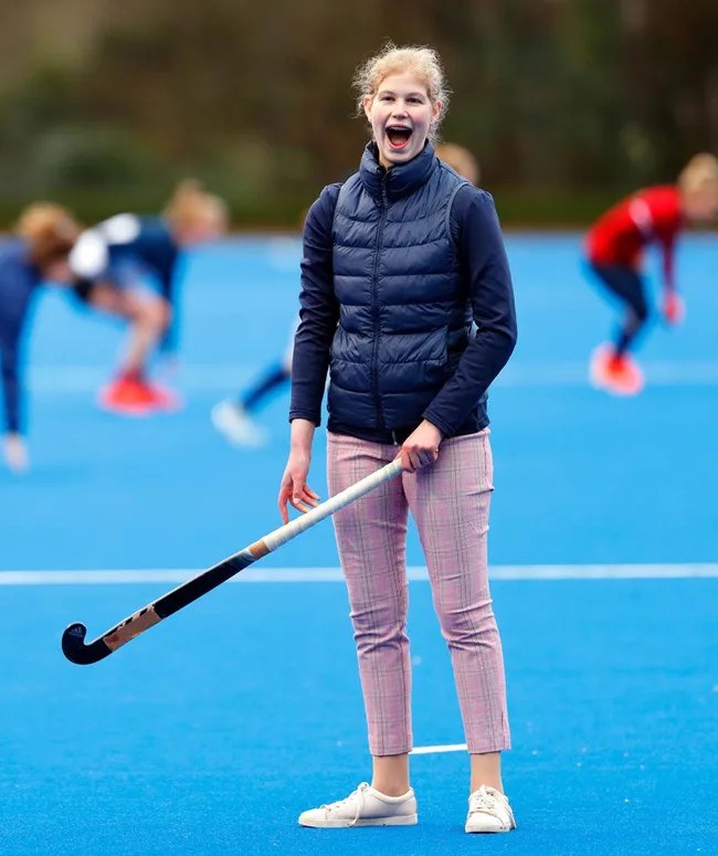 Girl holding a field hockey stick on a blue field, wearing a navy vest and pink pants, smiling brightly.
