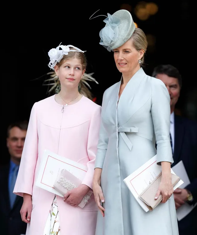 Two women in elegant pastel outfits and hats, holding programs, standing outdoors at a formal event.