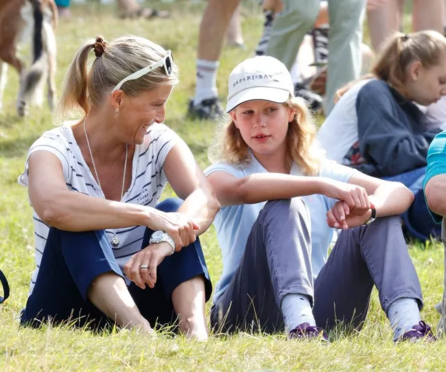 Two people sitting on grass, smiling and talking, outdoors with others in background. Casual attire, sunny day.