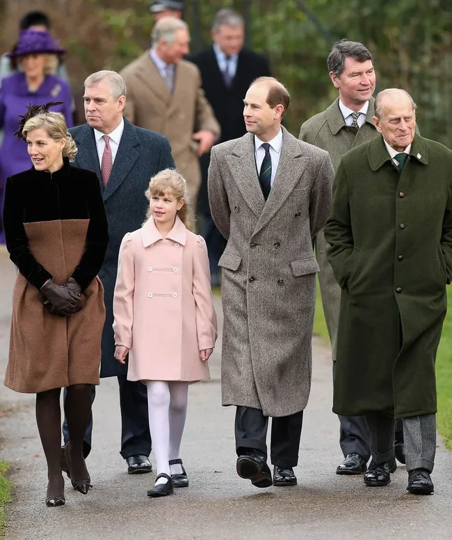 A group of people, including a girl in a pink coat, walk along a path wearing winter coats, with others in the background.