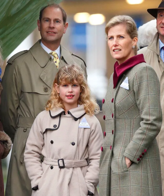 Three people in coats at an outdoor event, including a child in a beige coat and two adults in plaid and trench coats.