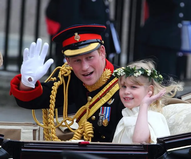 Two people waving from a carriage; one in military attire and a young girl with a flower crown.